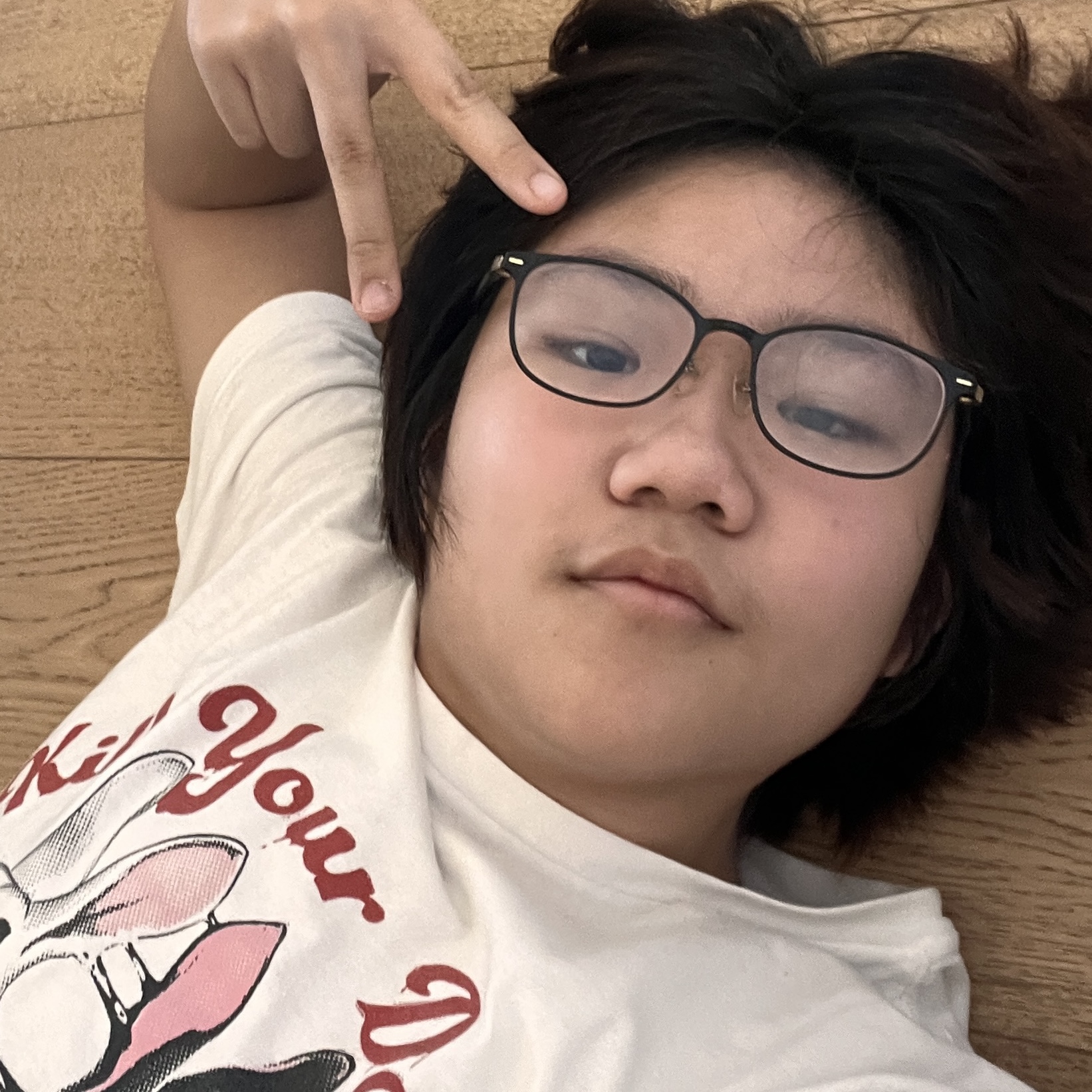 Selfie of an Asian teenager taken from a low angle. He has on glasses, and a T-shirt that says <...> 'your d', with a white rabbit and a matching black rabbit. He has black hair with brown ends, and is smiling at the camera, making a peace sign. He is lying on the floor.